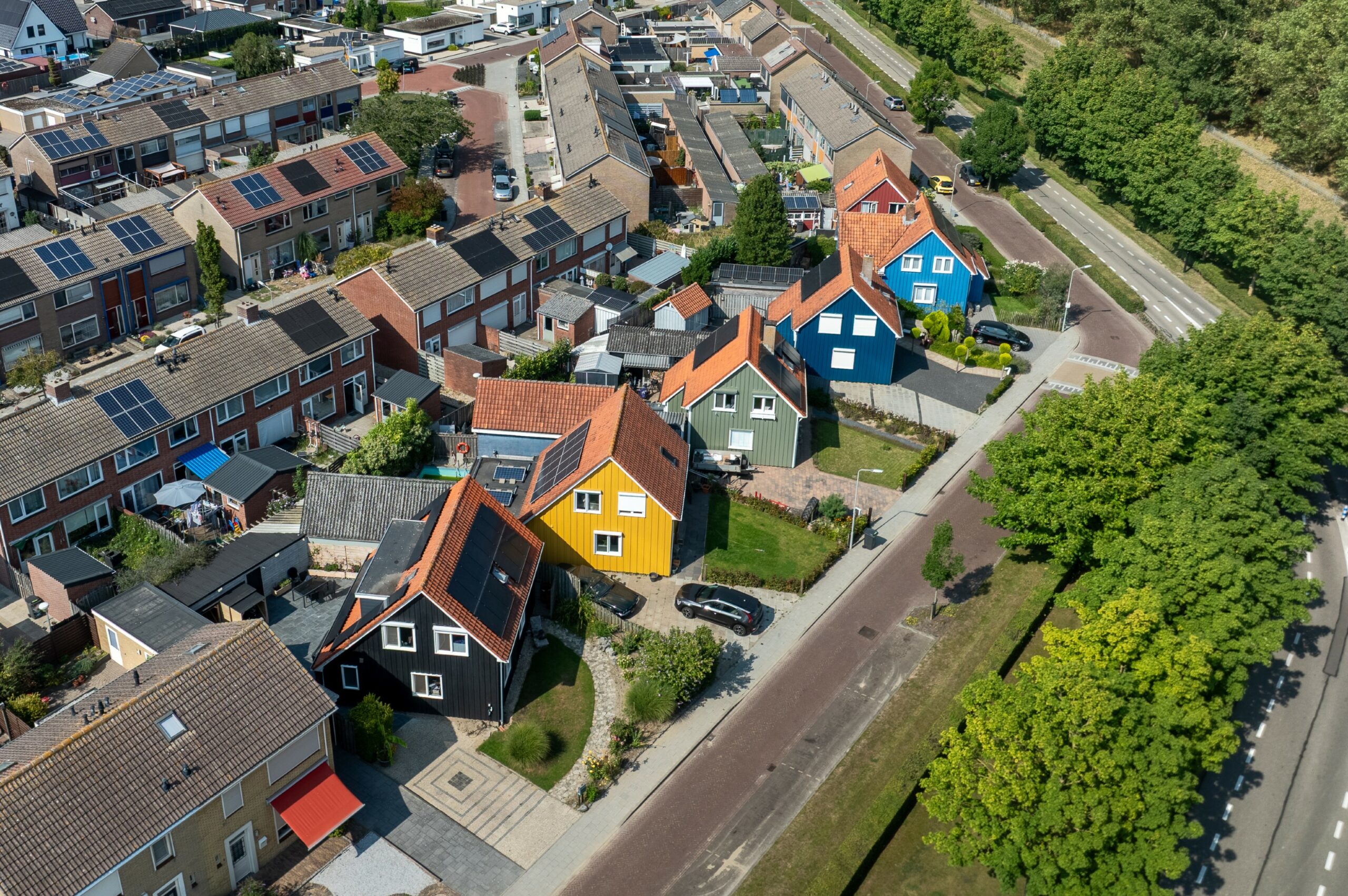 Flyfoto av et boligområde i Nederland med fargerike hus, inkludert freder 19 norske hus, solcellepaneler på takene, trær og parkerte biler langs gaten.