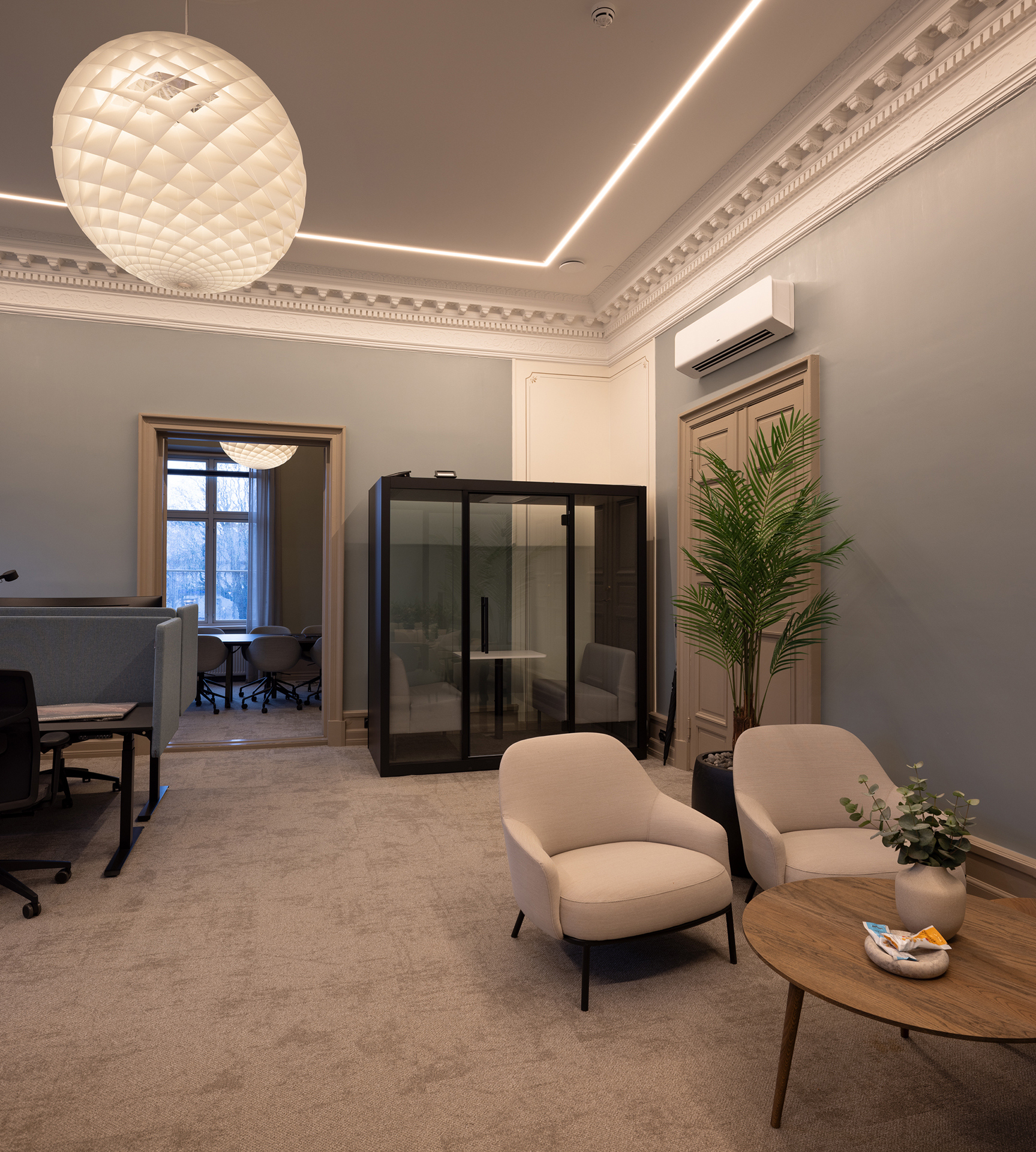 Moderne kontor med to lenestoler, et rundt salongbord, en potteplante, en telefonboks i glass og arbeidsstasjoner nær et stort vindu under dekorative taklamper.