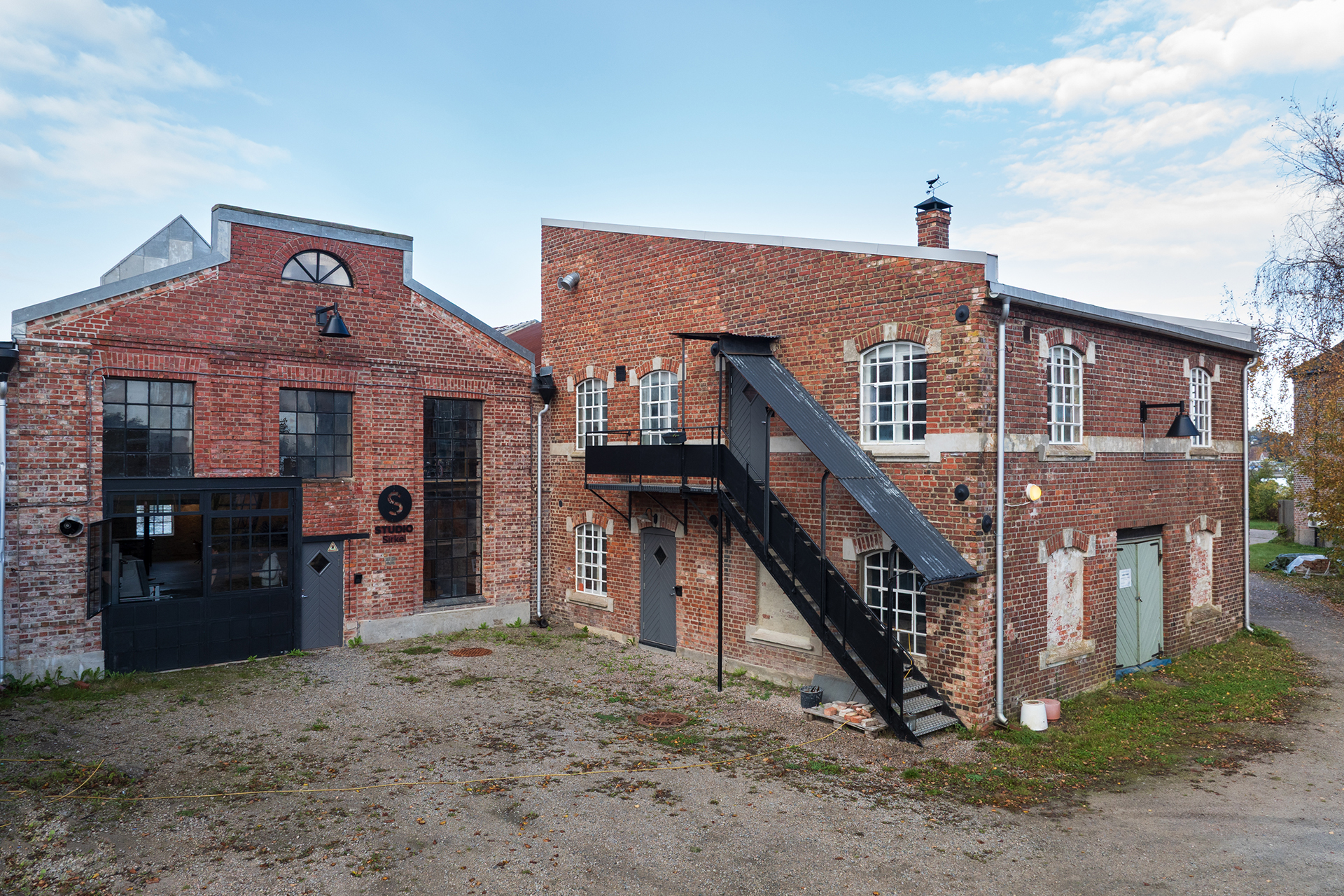 Sirkulærøkonomi i praksis 15 En industribygning i rød teglstein med store vinduer, metalldører og en utvendig svart metalltrapp, plassert på en gruset tomt under en delvis overskyet himmel.