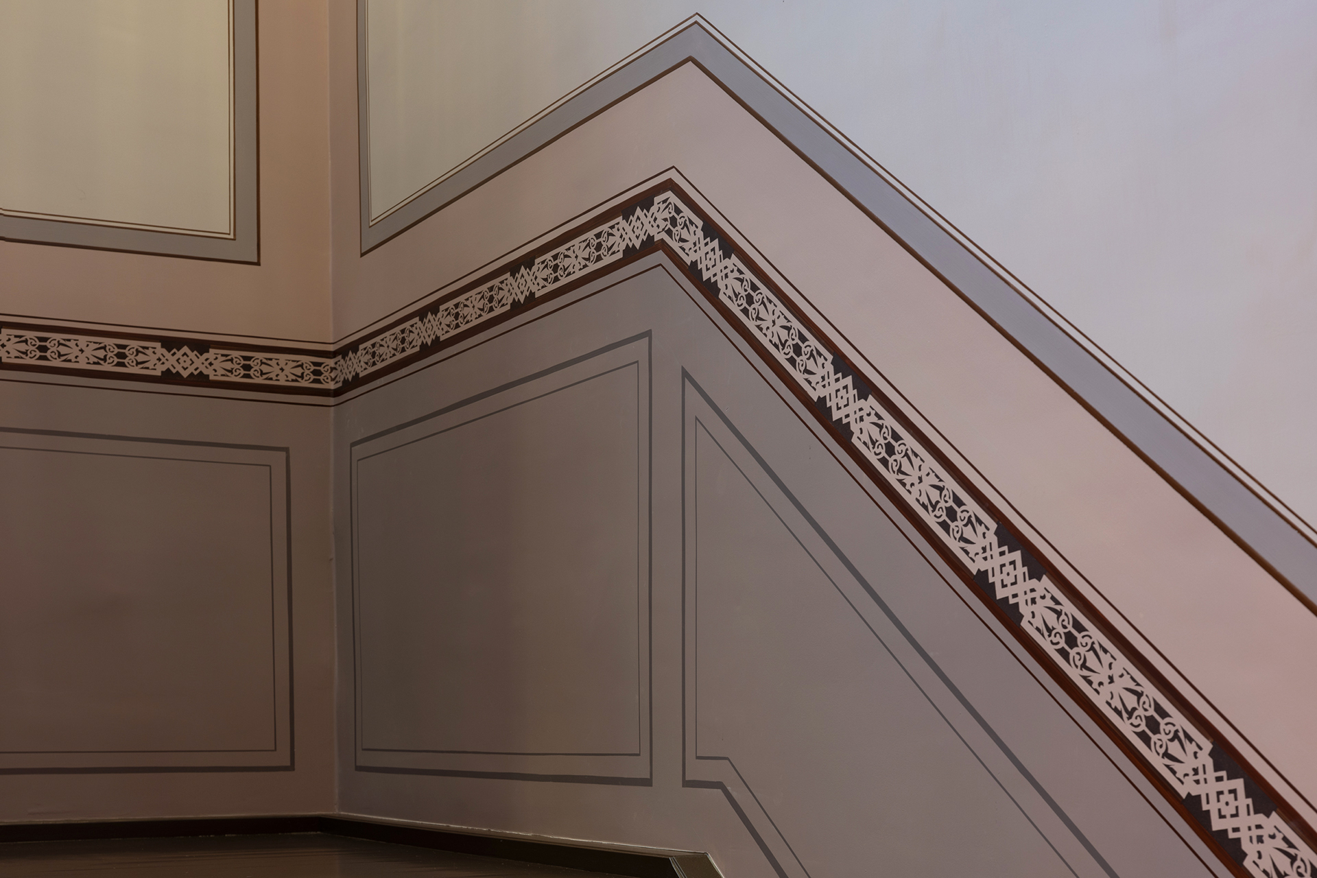 Innervegg med geometrisk dekorlist og mønstret listverk som følger vinkelen til en trapp, malt i beige og brune nyanser.