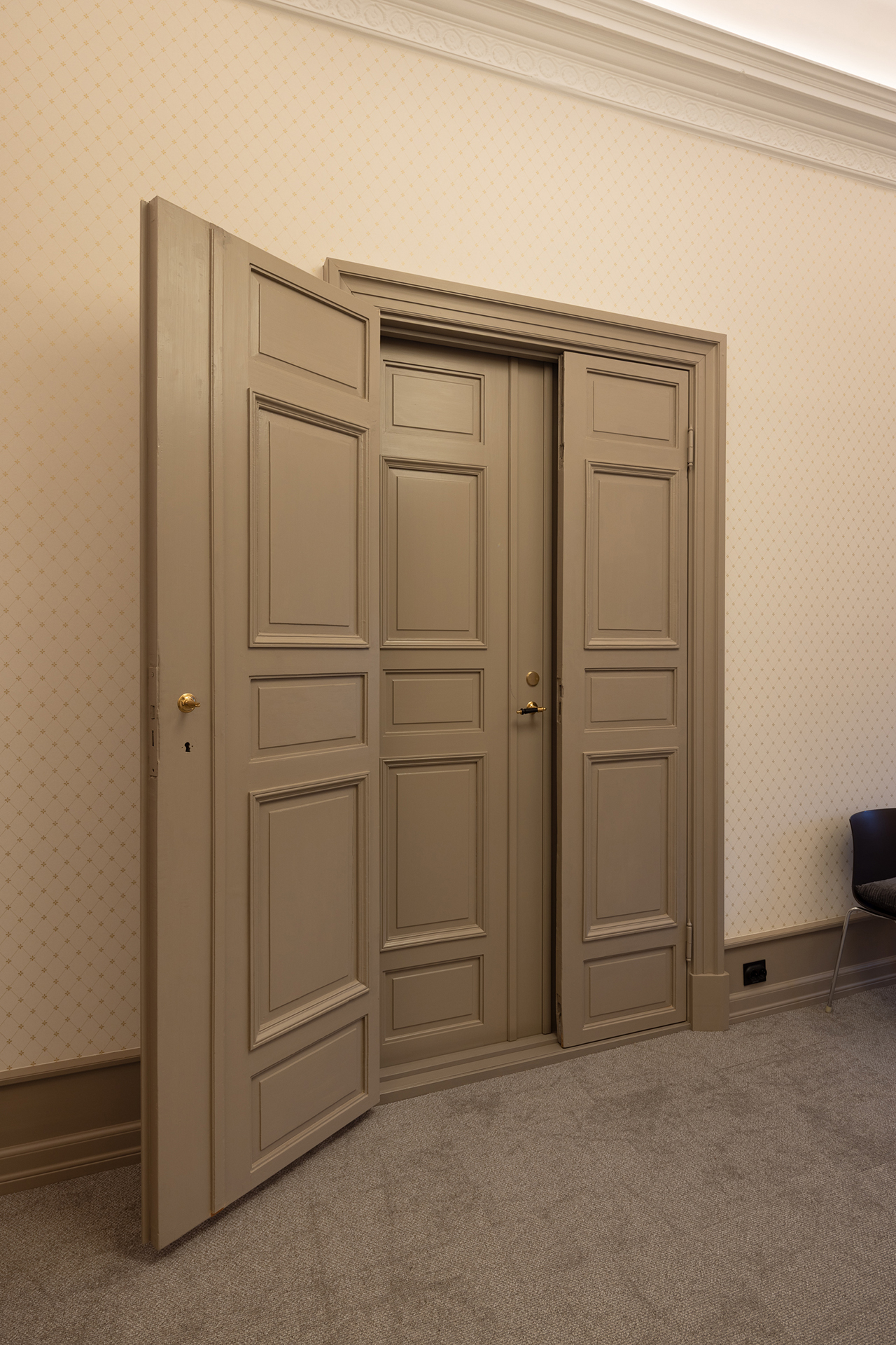 En liten dør i midten flankeres av to større, delvis åpne paneler i en beige vegg. En mørk stol står til høyre på et teppebelagt gulv.