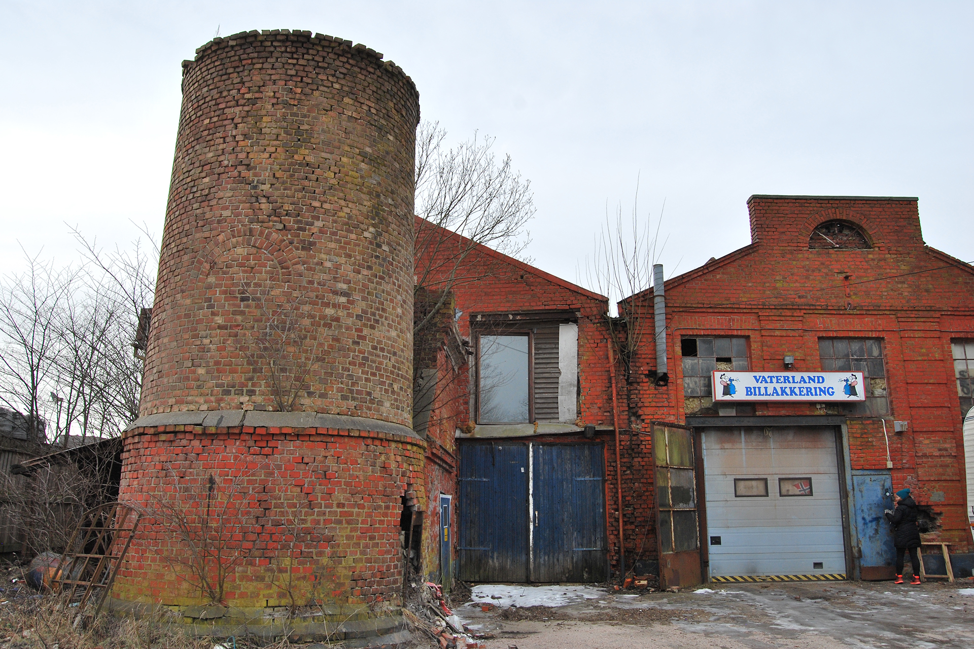 Industribygning i rød teglstein med et stort sylindrisk tårn, blå garasjeporter og et skilt med Vaterland Billakering. Noen vinduer er knust, og området ser forsømt ut.