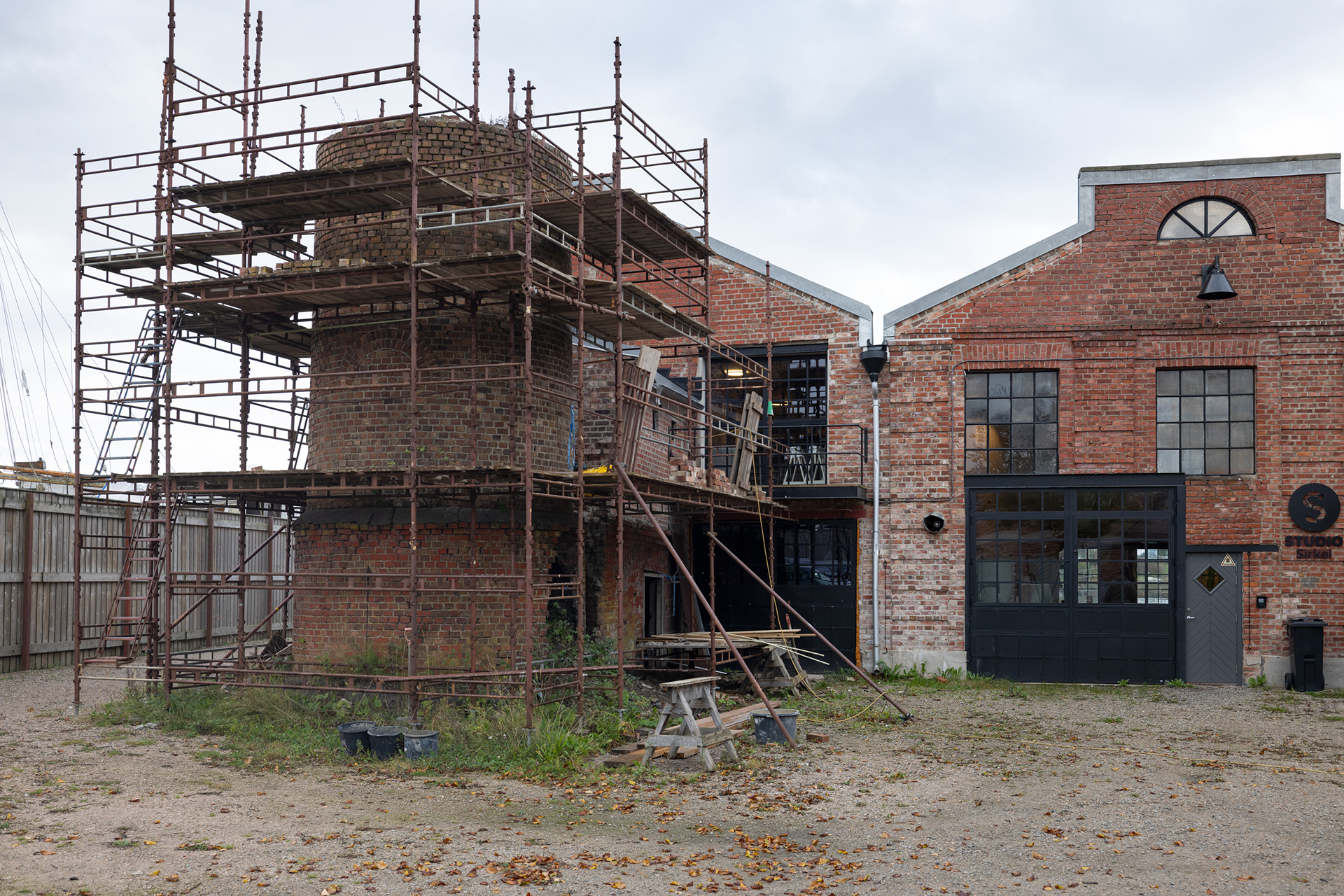 En mursteinsbygning under oppussing med stillaser av metall omkranser en sylinderformet struktur utenfor. Området er ryddet, med noen byggematerialer og et piknikbord i nærheten.