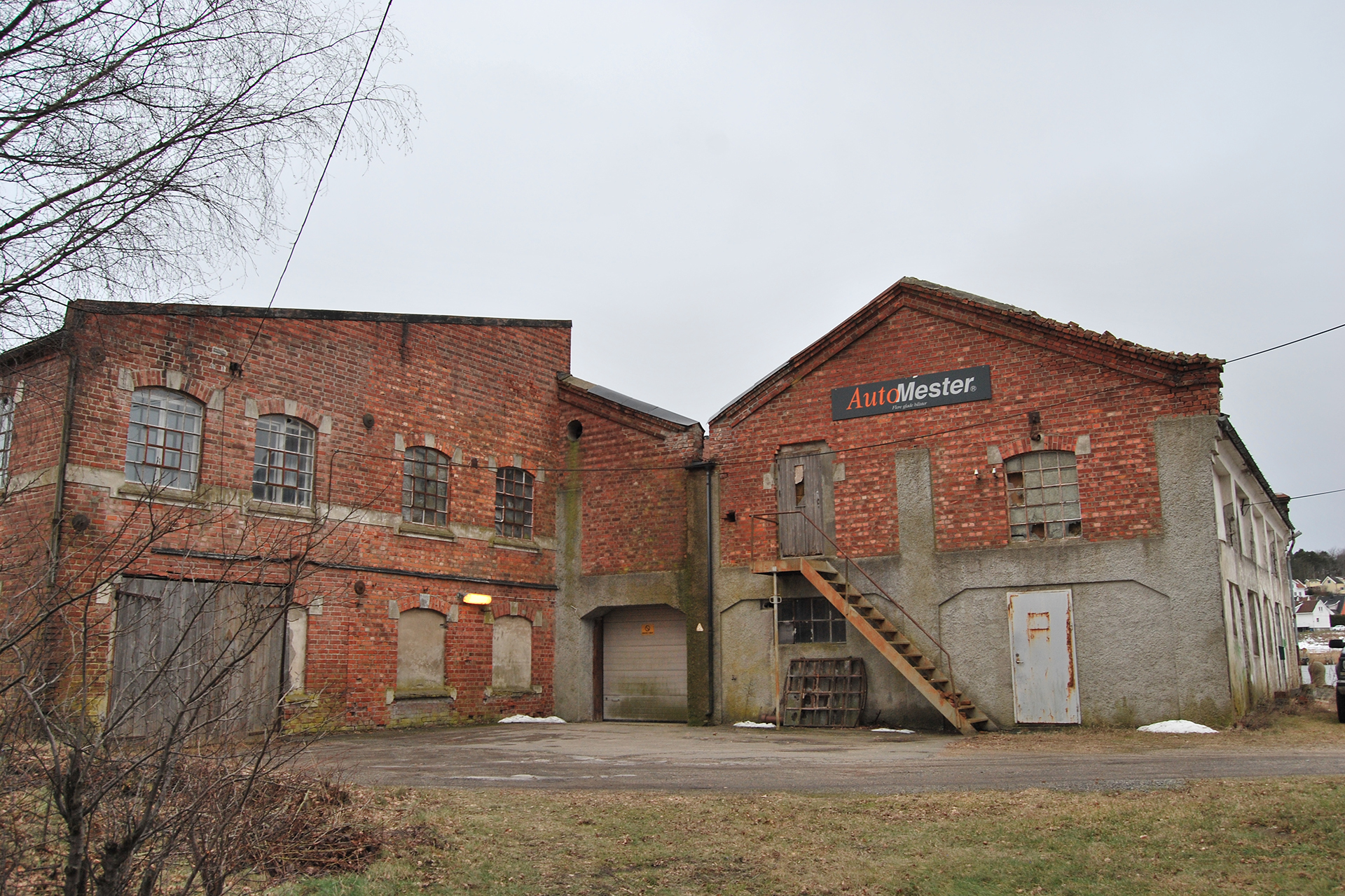 En industribygning i murstein og betong med et skilt med teksten AutoMester over en tretrapp, omgitt av løvløse trær og snøflekker.