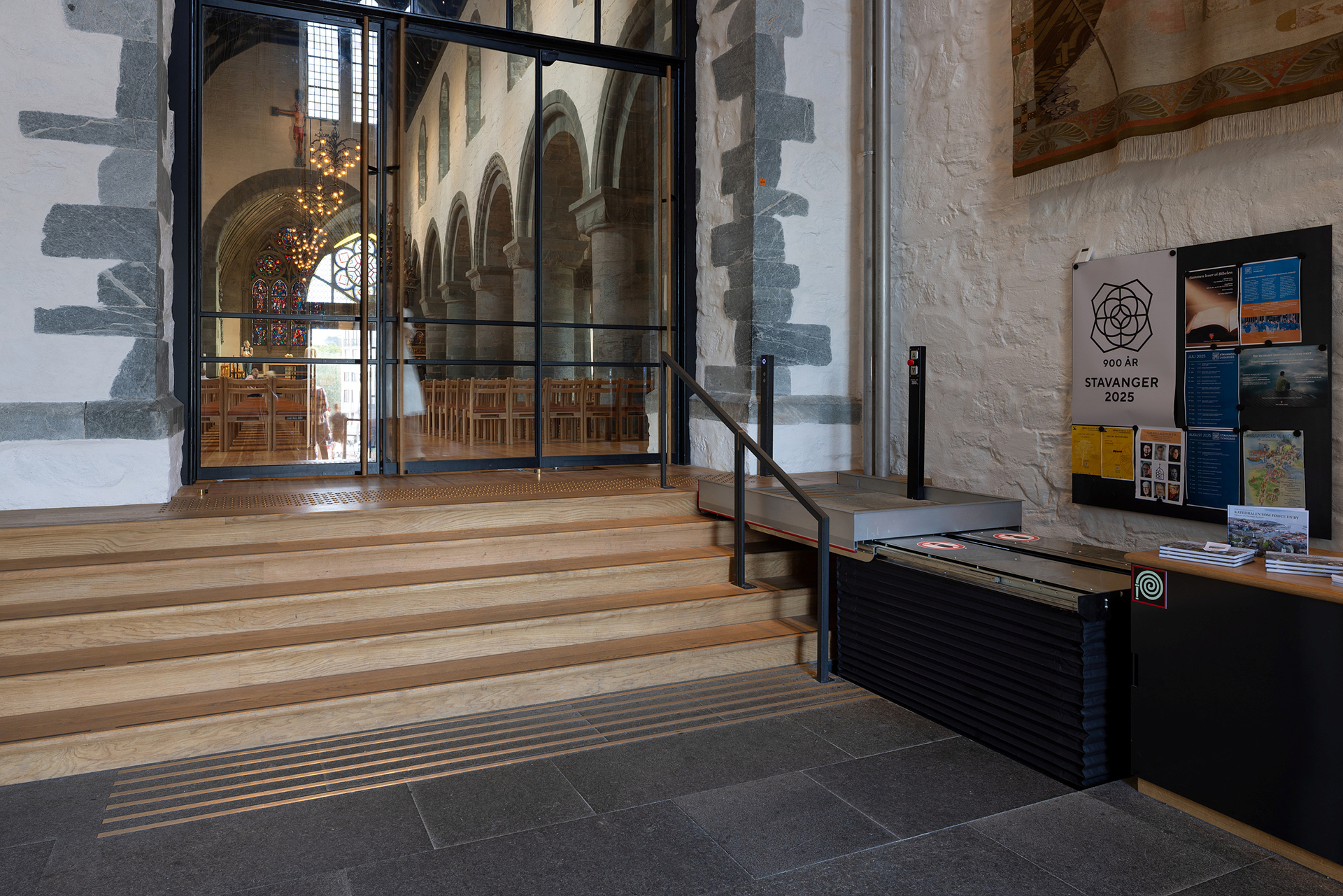 Moderne tilgjengelegheit i mellomalderkyrkje 9 En tretrapp og en plattformheis gir tilgang til et kirkerom med steinbuer og sitteplasser i tre; informasjonsbrosjyrer ligger utstilt til høyre.