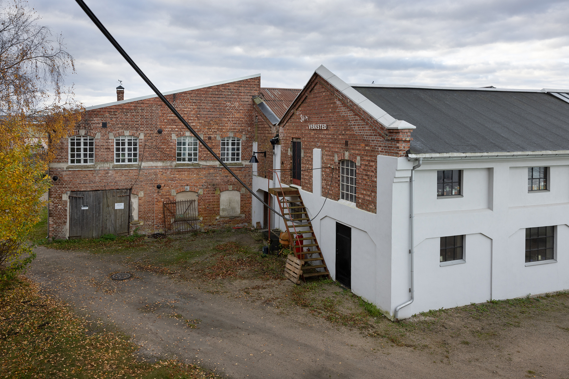 En industribygning i murstein og hvitt med en utvendig tretrapp, omgitt av en grusvei og høsttrær under en overskyet himmel.