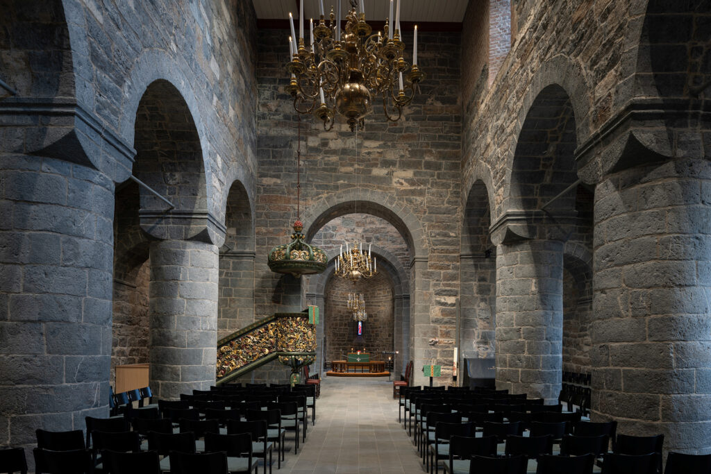 Interiør av stein i en kirke med buede søyler, rekker av svarte stoler, utsmykkede lysekroner i gull, en dekorert prekestol og et alter foran.