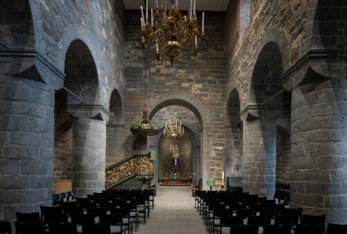 Interiør av stein i en kirke med buede søyler, rekker av svarte stoler, utsmykkede lysekroner i gull, en dekorert prekestol og et alter foran.