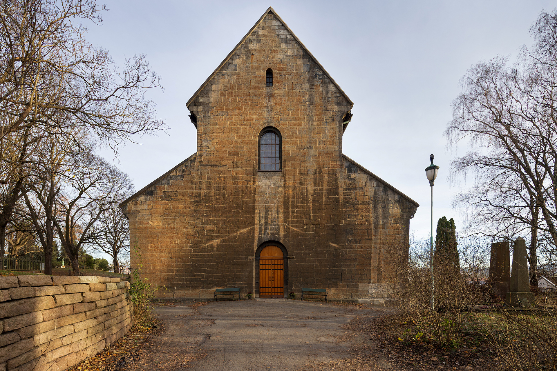 En steinkirke med et høyt, spisset tak, buede vinduer og en tredør, omgitt av nakne trær og en steinmur på en overskyet dag.