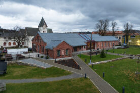En rød mursteinsbygning med metalltak står ved siden av en kirke med et høyt kirketårn, omgitt av stier, gress, benker og løvløse trær under en overskyet himmel.