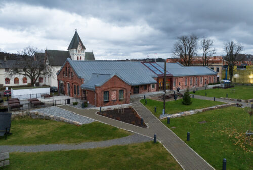 Eksempelsamling 3 En rød mursteinsbygning med metalltak står ved siden av en kirke med et høyt kirketårn, omgitt av stier, gress, benker og løvløse trær under en overskyet himmel.