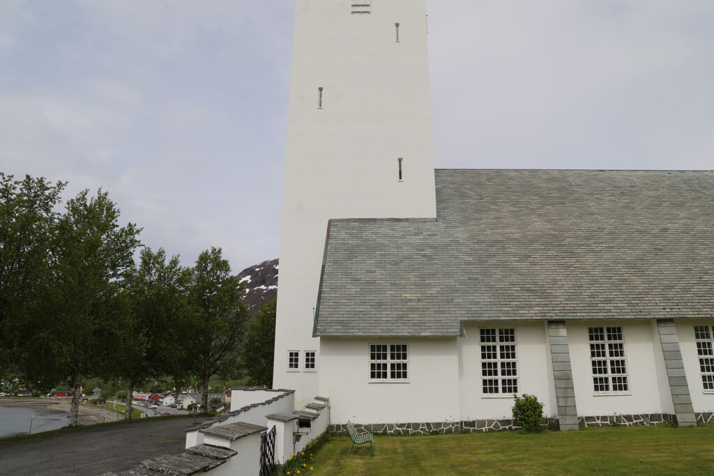 Hvit kirke med et høyt, rektangulært tårn og grått tak, plassert på en gressplen med trær og fjell i bakgrunnen.