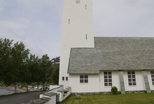 Hvit kirke med et høyt, rektangulært tårn og grått tak, plassert på en gressplen med trær og fjell i bakgrunnen.