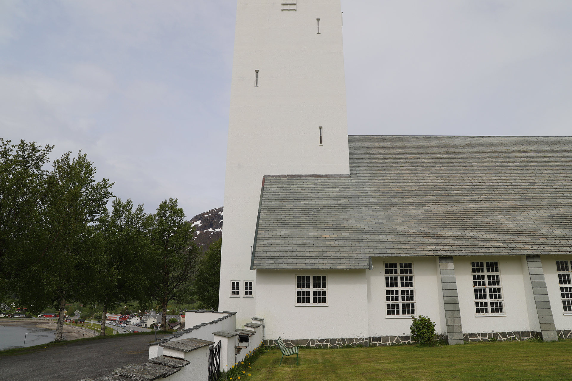 Hvit kirke med et høyt, rektangulært tårn og grått tak, plassert på en gressplen med trær og fjell i bakgrunnen.