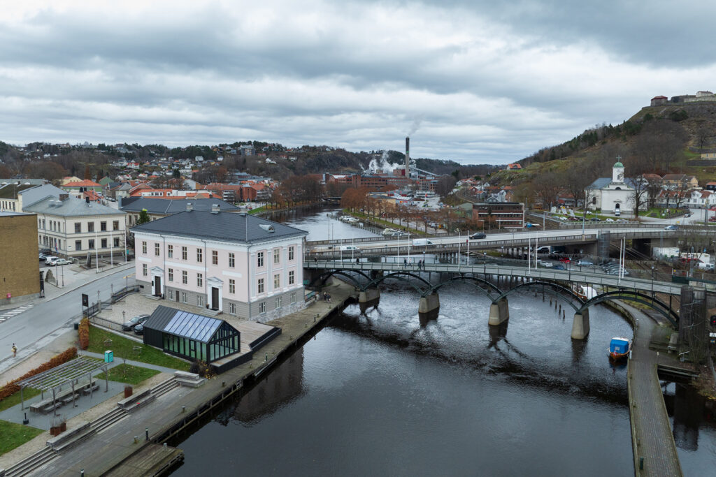 En by ved elvebredden med en bro over vannet, omgitt av bygninger, trær og en høyde med en festning under en overskyet himmel.
