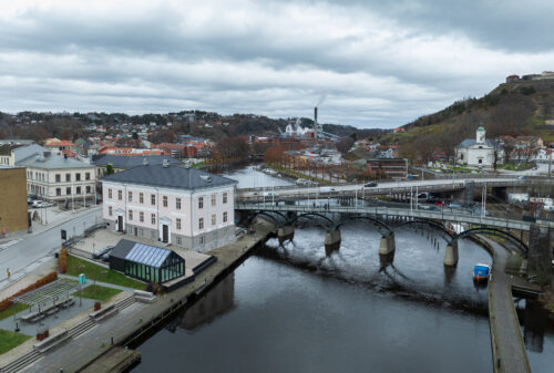Eksempelsamling 6 En by ved elvebredden med en bro over vannet, omgitt av bygninger, trær og en høyde med en festning under en overskyet himmel.