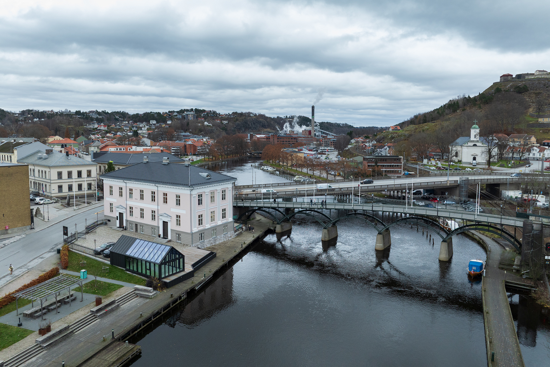 En by ved elvebredden med en bro over vannet, omgitt av bygninger, trær og en høyde med en festning under en overskyet himmel.