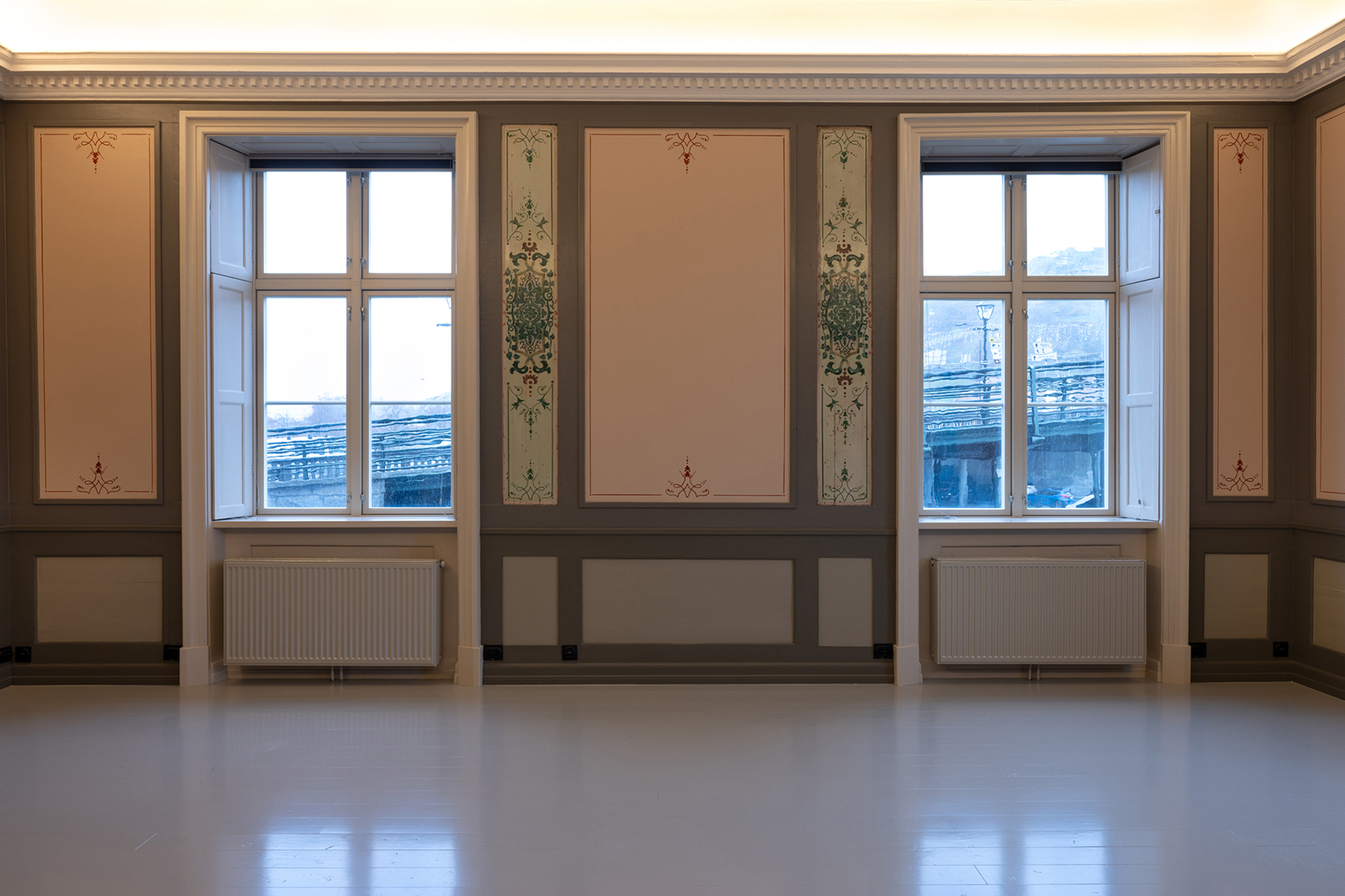 Et rom med tre store vinduer, beige og brune panelvegger, dekorative grønne detaljer og et blankpolert, lyst gulv.