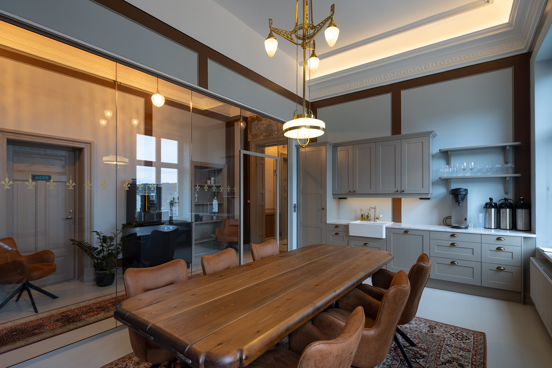 Et moderne møterom med trebord, åtte brune stoler, en kjøkkenkrok og glassvegger som skiller det fra et tilstøtende kontorlokale.