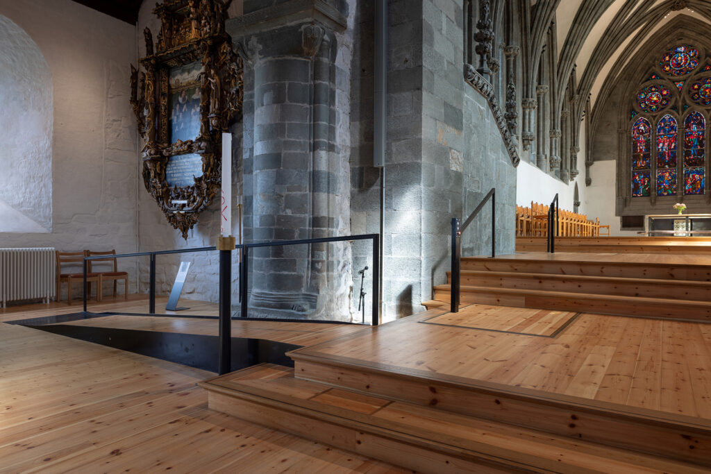 Interiør i en historisk kirke med steinvegger, glassmalerier, tregulv, trapper og en moderne rampe med glassrekkverk som fører opp til alterområdet.