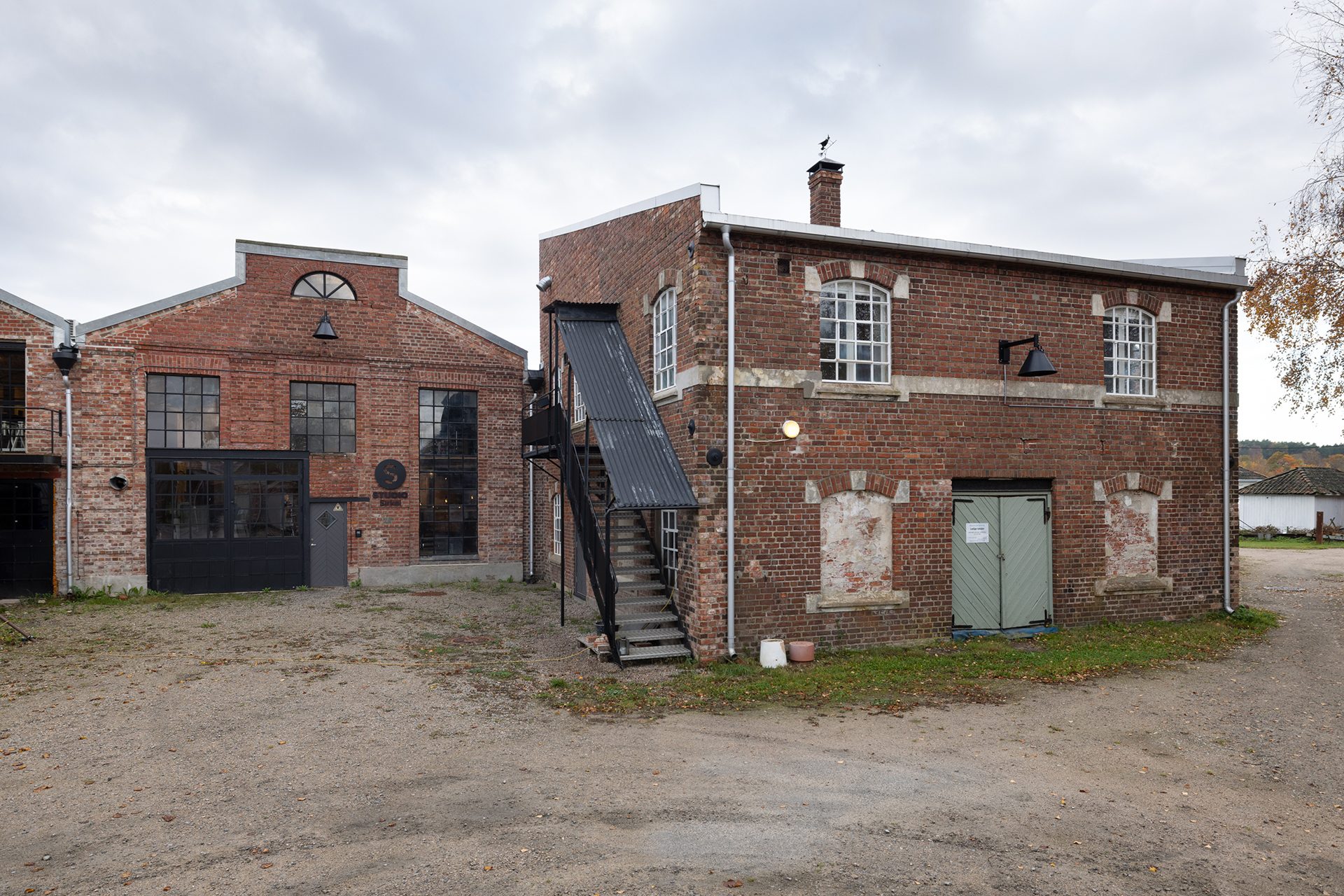 To industribygninger i murstein ligger side om side på en gruset tomt, der den ene har en utvendig trapp og den andre store industrivinduer og doble dører.