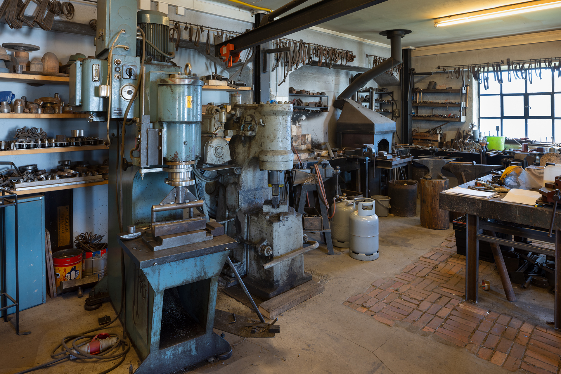 Sirkulærøkonomi i praksis 8 Et verksted med industrimaskiner, bensintanker, verktøy på hyller, en smie og en arbeidsbenk, med naturlig lys fra et vindu til høyre.