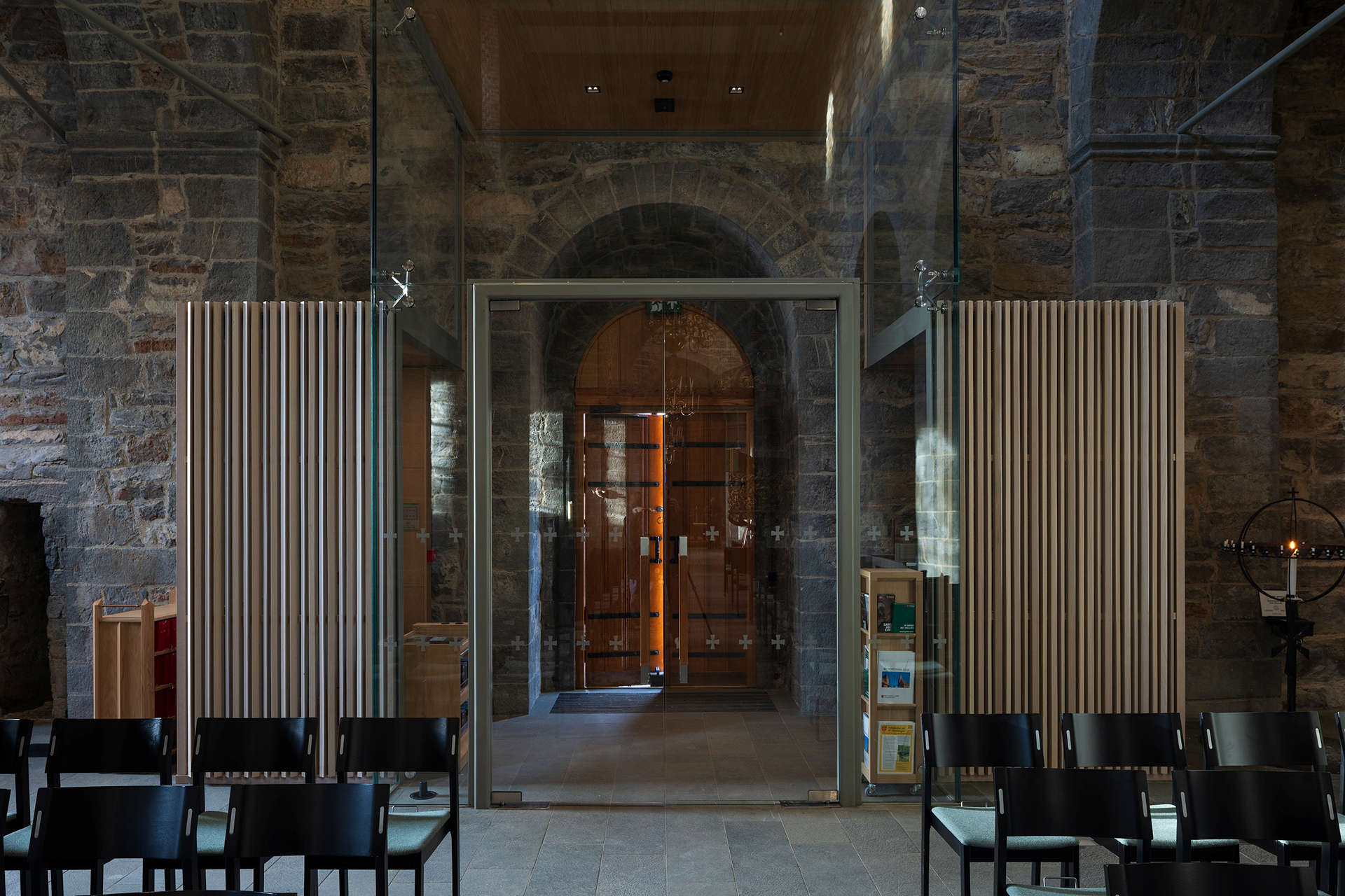 Moderne inngangsparti i glass med metalldører innenfor en steinmur, flankert av vertikale trelister; svarte stoler står på rekke og rad i forgrunnen.