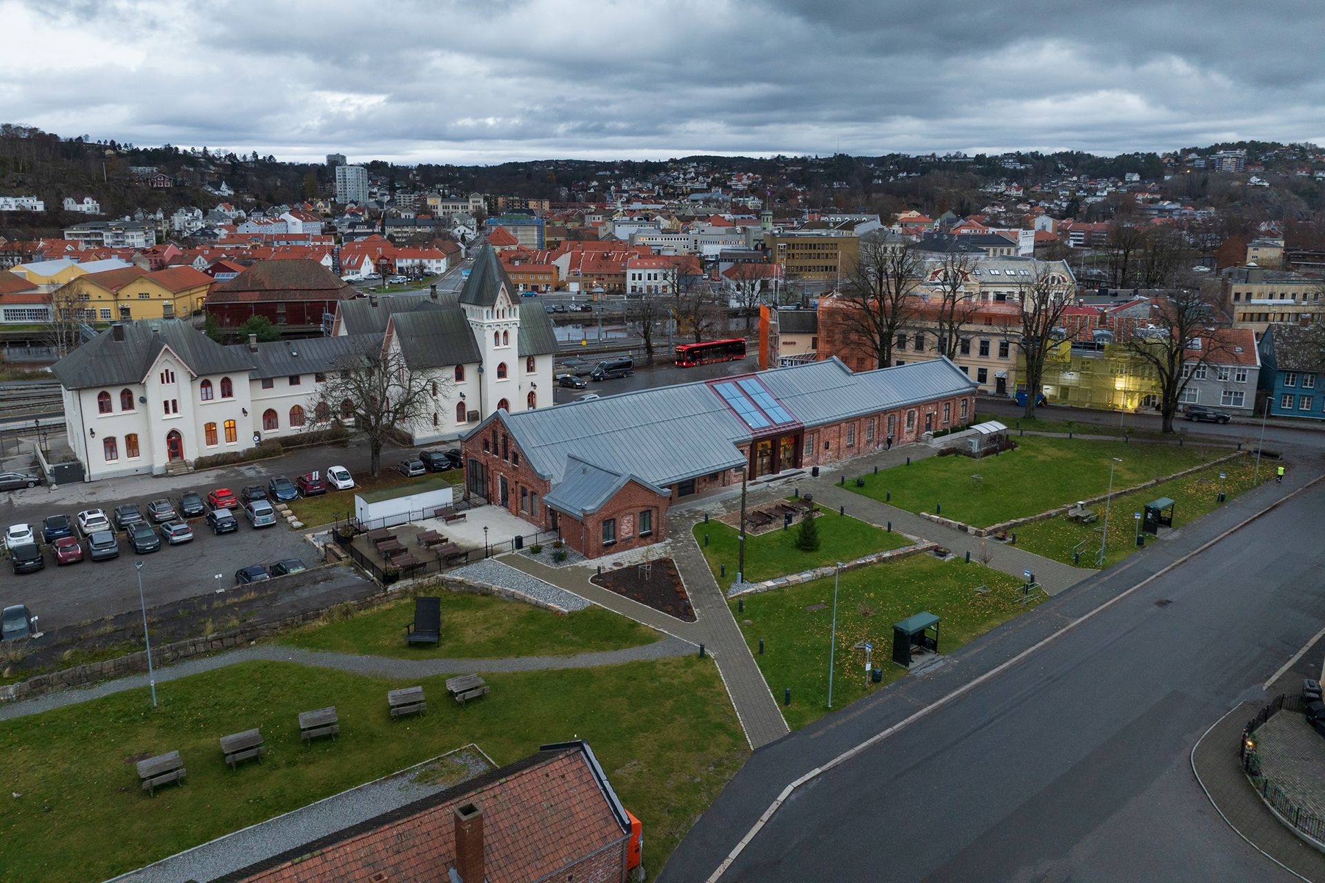 På rett spor i Byparken 6 Et luftfoto av en historisk mursteinsbygning med glasstak og en hvit kirkelignende struktur, omgitt av grønne plener og benker, med en by og en overskyet himmel i bakgrunnen.