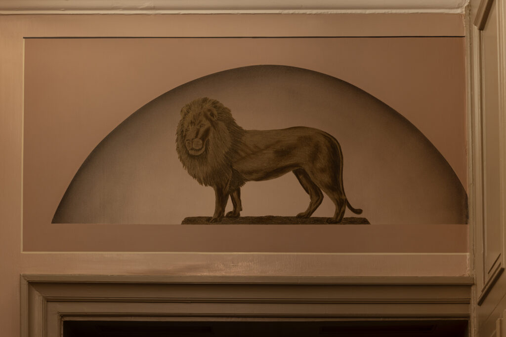Et sepiatonet veggmaleri av en løve som står på en stein, innrammet i en bueform over en døråpning innendørs.