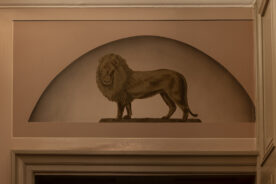 Et sepiatonet veggmaleri av en løve som står på en stein, innrammet i en bueform over en døråpning innendørs.