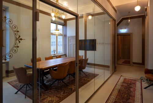 Eksempelsamling 5 Et moderne konferanserom med et langt trebord, brune stoler, glassvegger og innredning i vintage-stil, sett fra en gang med tepper og varm belysning.