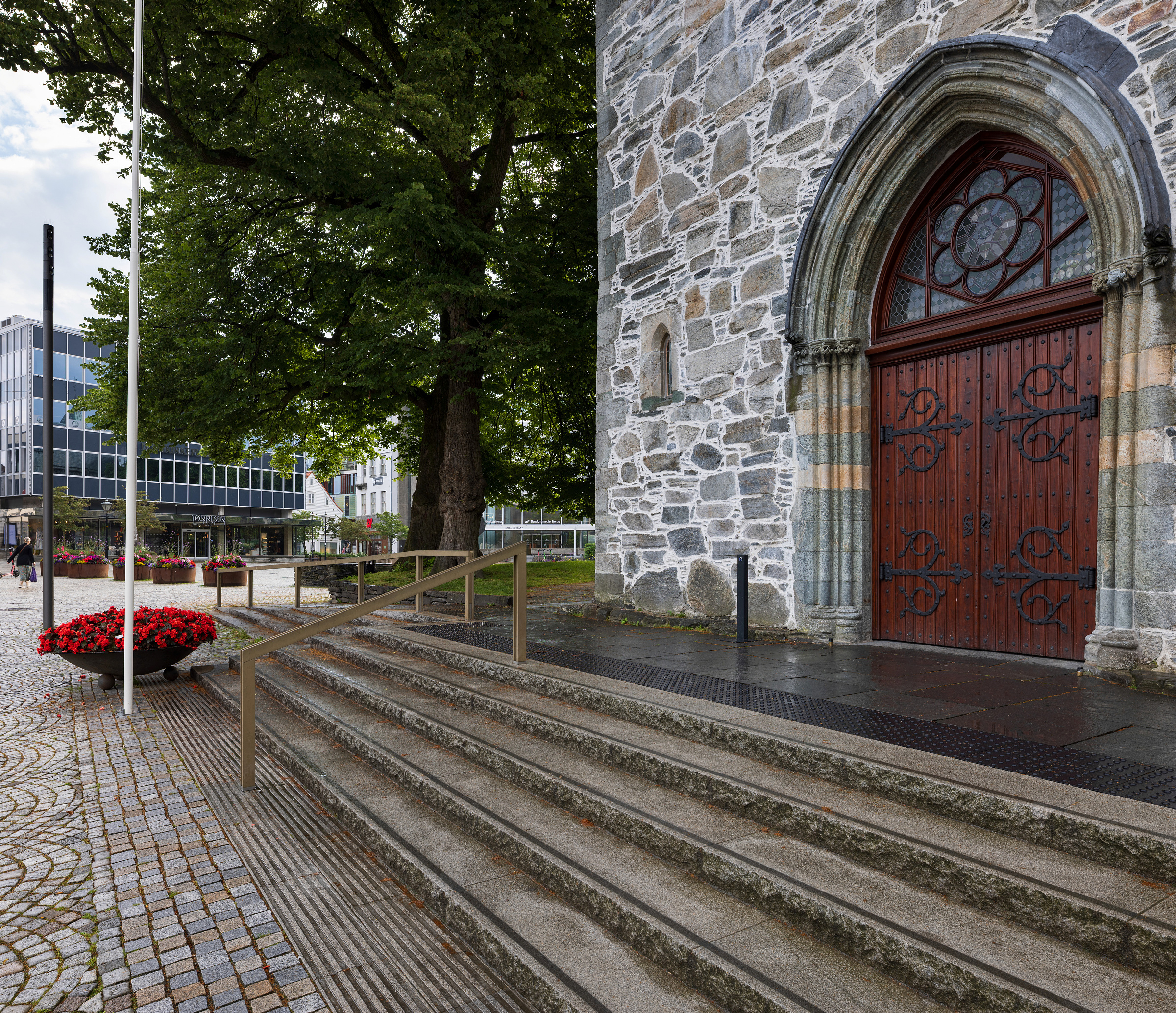Moderne tilgjengelegheit i mellomalderkyrkje 6 Inngangsparti i stein med utsmykkede tredører, trapper, rekkverk og trær i nærheten. Røde blomster er plassert i store krukker på brosteinsbelagte gangveier.