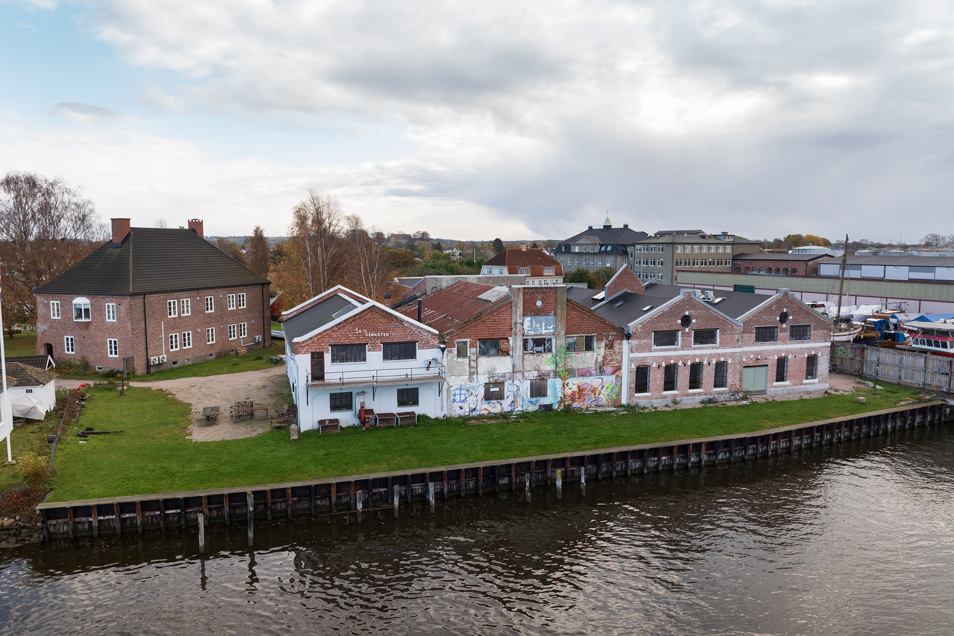En rekke gamle mursteinsbygninger, noen med graffiti og synlig slitasje, står langs et gressletteområde ved en elv under en overskyet himmel.