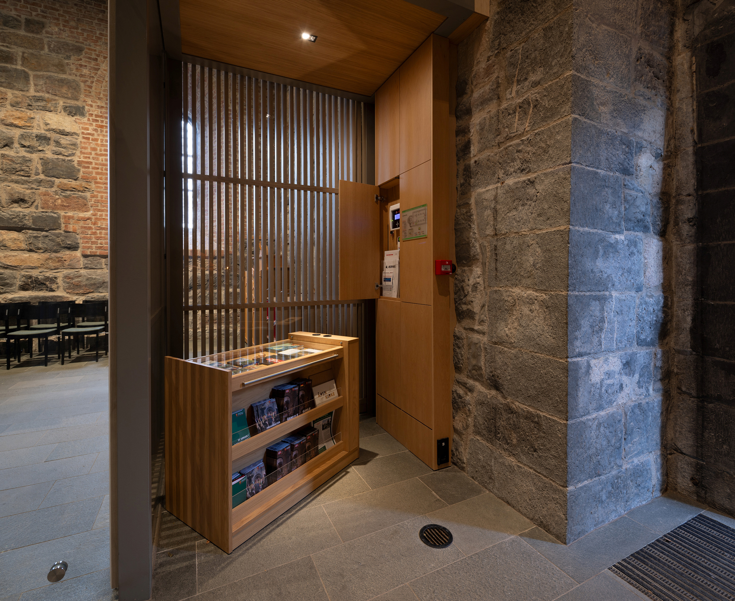 En informasjonskiosk i tre med brosjyrer står ved en steinvegg i et moderne interiør med grått flisgulv og dempet belysning.