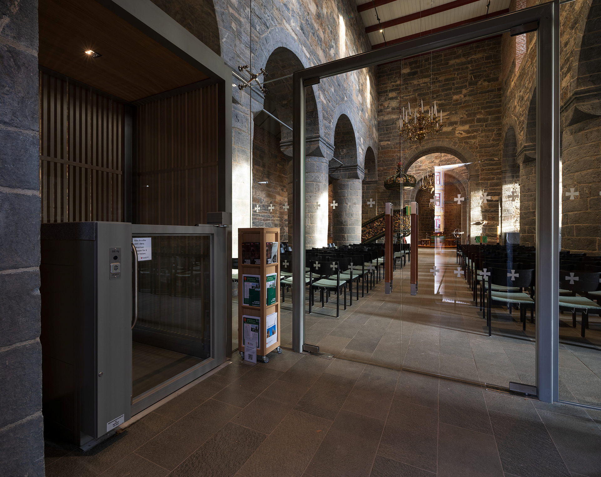 En moderne heis og en brosjyre om universell utforming står ved inngangen til en steinkirke med buede søyler, glassdører og rader med sitteplasser.