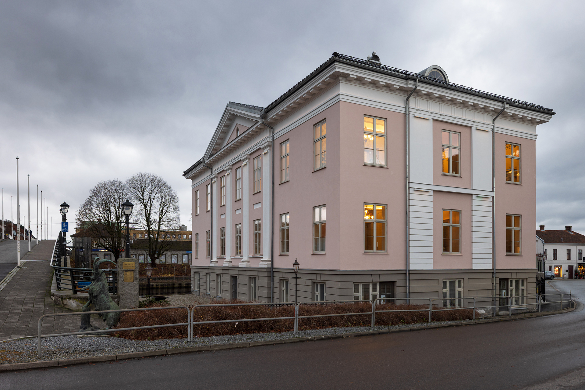 En blekrosa, toetasjes nyklassisistisk bygning med opplyste vinduer står på et gatehjørne, ved siden av en gangvei med lyktestolper og løvløse trær under en overskyet himmel.