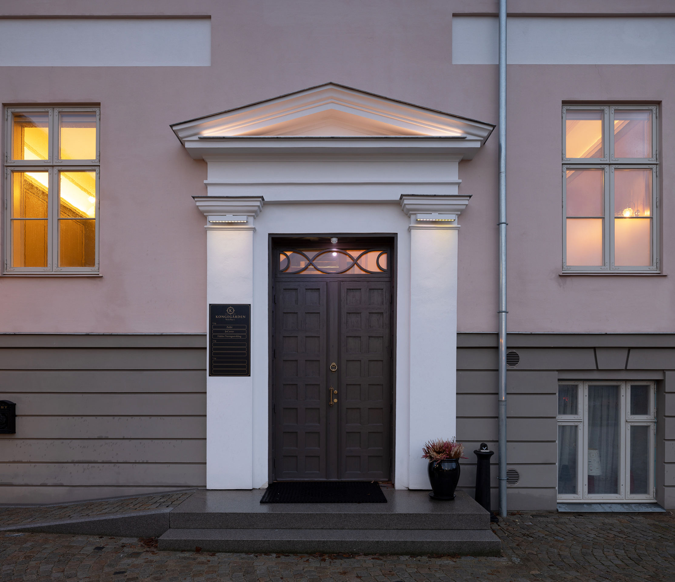 En grå inngangsdør med dekorativ listverk er satt inn i en blekrosa og grå bygning, flankert av to vinduer og en liten trapp som fører opp til inngangspartiet.