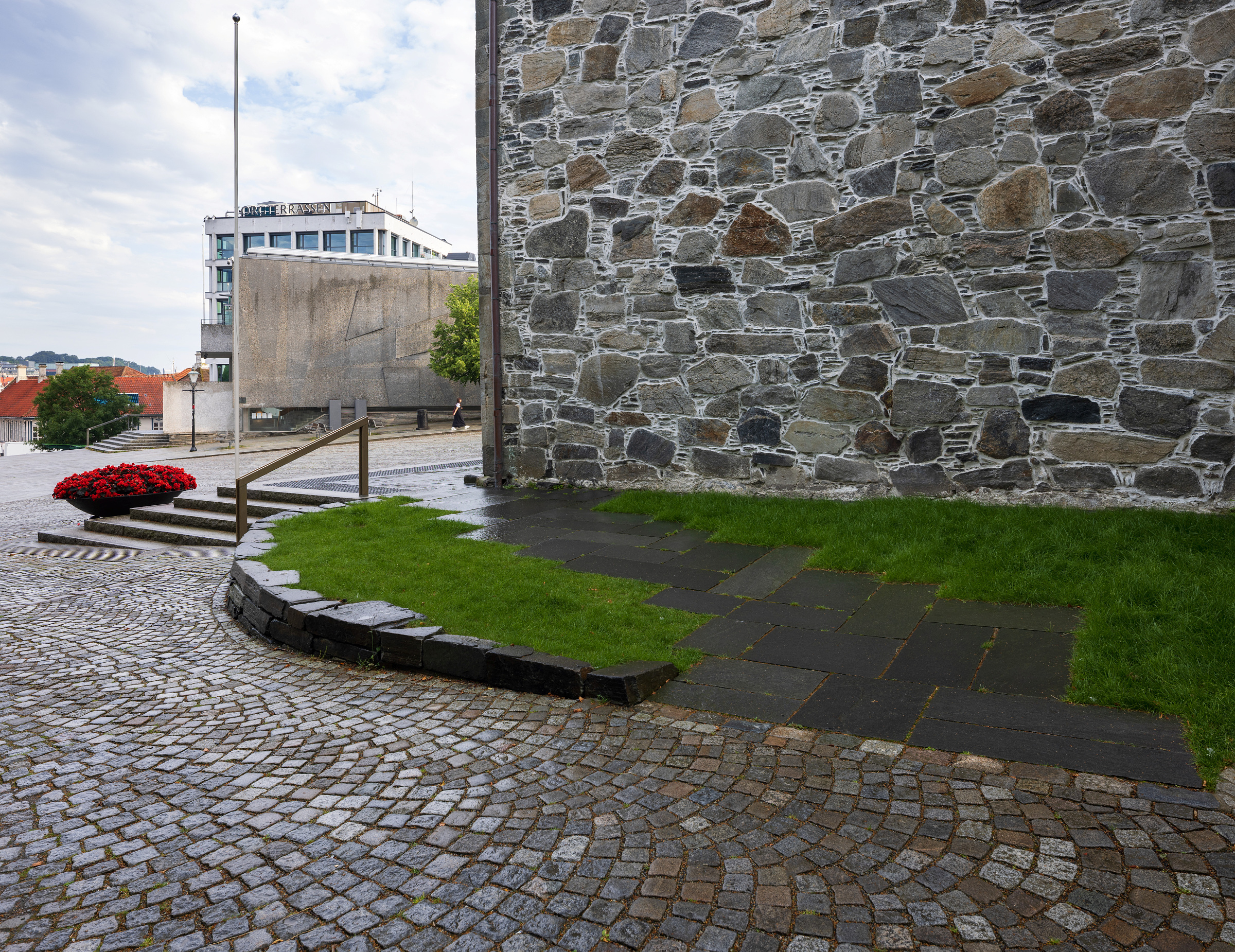 Moderne tilgjengelegheit i mellomalderkyrkje 7 En steinmur avgrenser et område med grønt gress og svarte fliser, omgitt av buet brostein og trappetrinn med et rødt blomsterarrangement i nærheten.