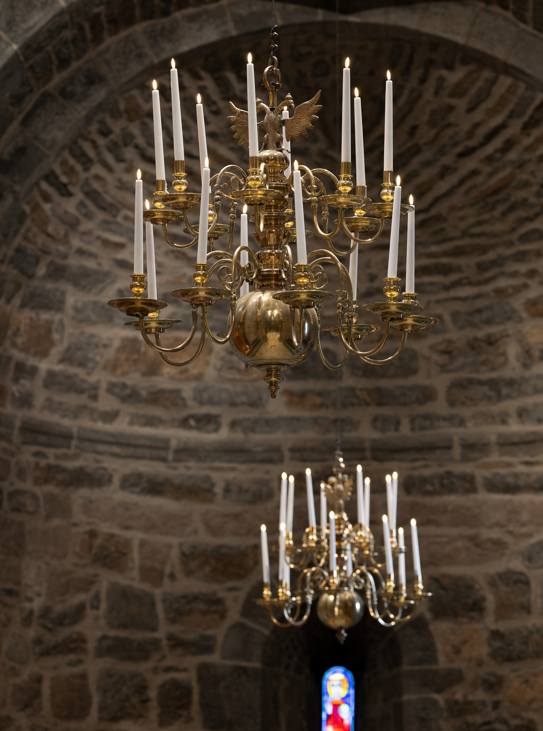 To utsmykkede lysekroner i messing med tente stearinlys henger fra et tak med steinbuer, og i bakgrunnen skimtes et glassmaleri.