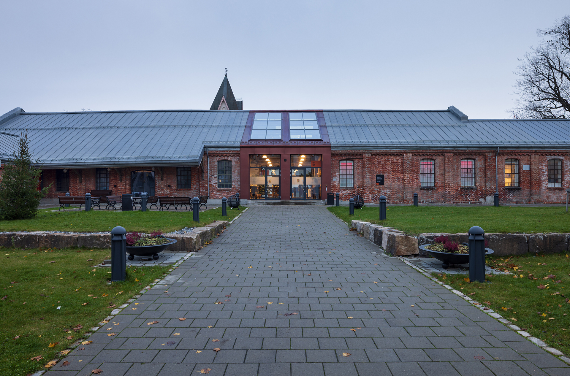 På rett spor i Byparken 10 En mursteinsbygning med spiss metalltak og glassinngang, omgitt av gress, benker og asfaltert gangvei på en overskyet dag.