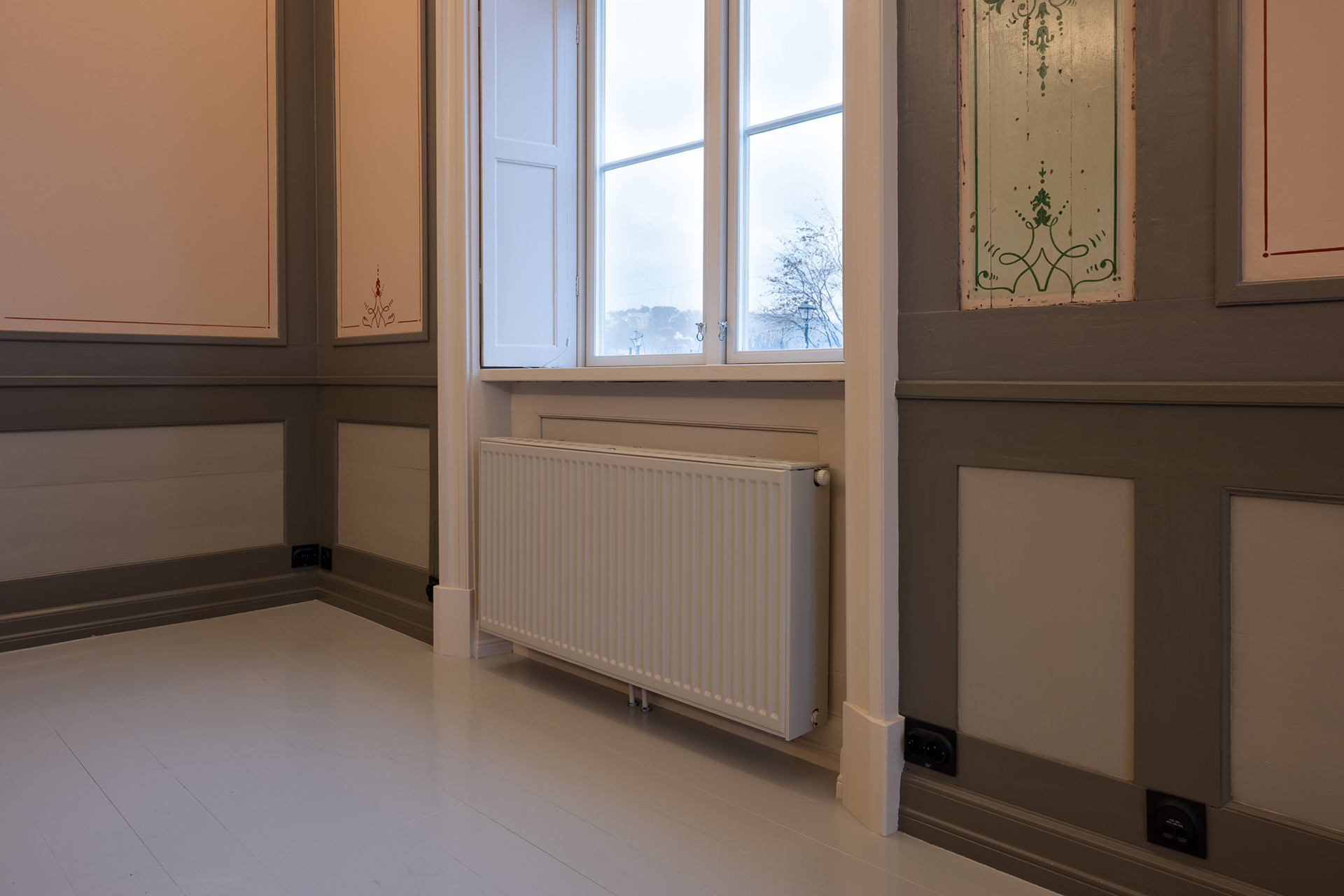 En hvit radiator er montert under et vindu med delvis åpne skodder i et rom med brune og beige panelvegger og lyst gulv.