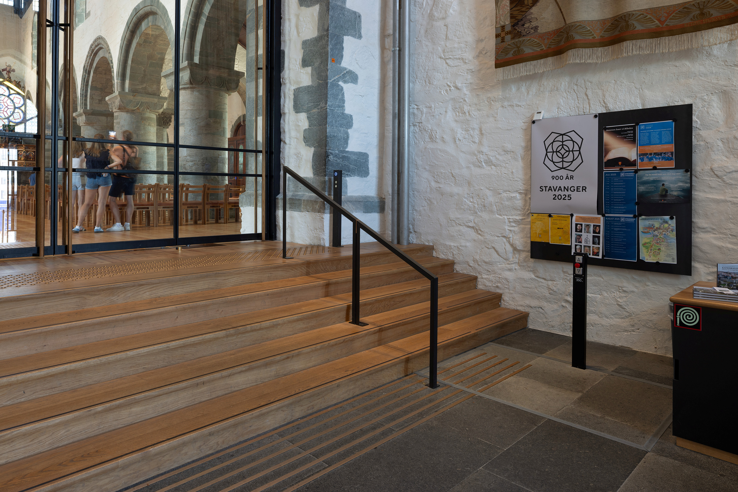 Moderne tilgjengelegheit i mellomalderkyrkje 8 Interiør i en historisk steinbygning med tretrapper, et svart rekkverk, informasjonsskjermer på veggen og mennesker som er synlige gjennom en glassdør i bakgrunnen.