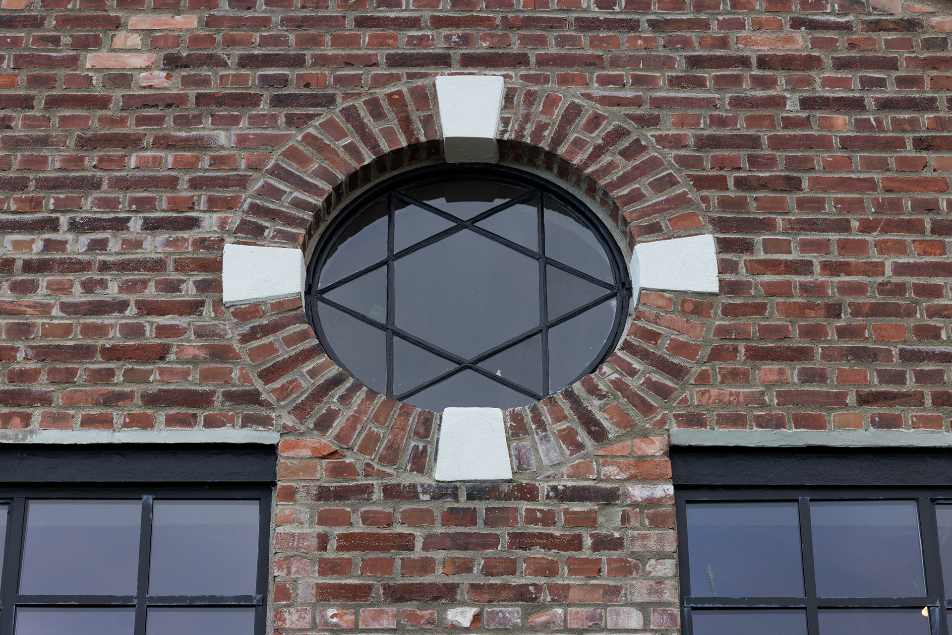 Sirkulært vindu med hvite teglsteinsdetaljer og svart rutenettmønster på en rød murvegg, over rektangulære vinduer med svarte rammer.