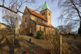 En steinkirke med et klokketårn med grønt tak står på en kirkegård med gravsteiner og nakne trær på en klar dag.