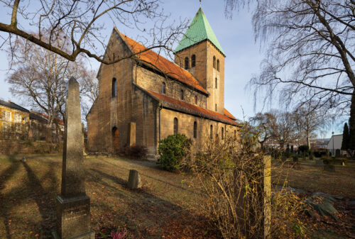 En steinkirke med et klokketårn med grønt tak står på en kirkegård med gravsteiner og nakne trær på en klar dag.