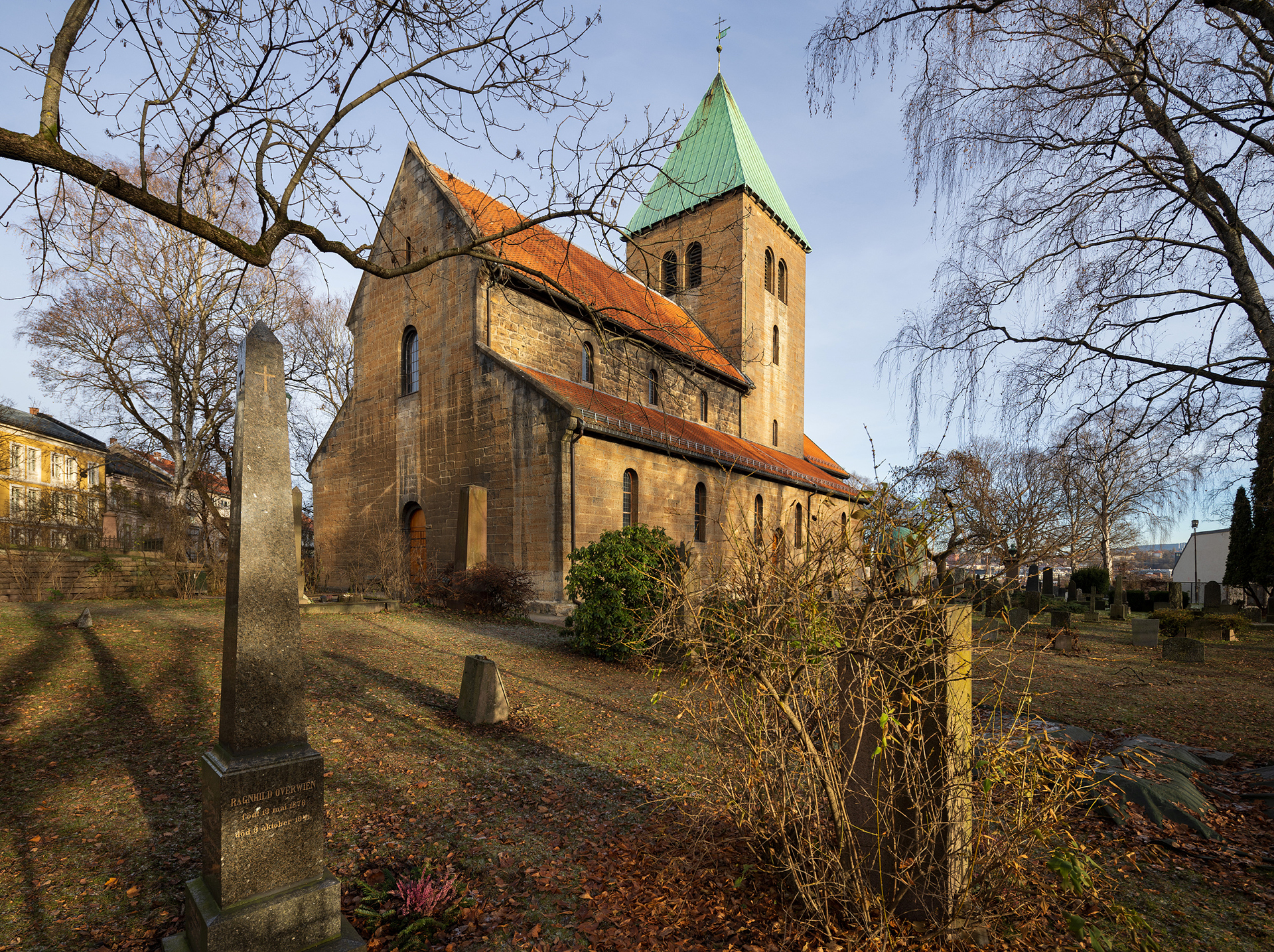 En steinkirke med et klokketårn med grønt tak står på en kirkegård med gravsteiner og nakne trær på en klar dag.