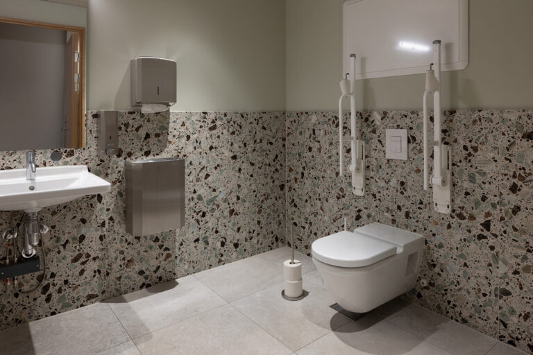 Tilgjengelig toalett med veggmontert vask, støttestativ ved siden av vegghengt toalett, toalettpapirholder og flislagte vegger og gulv.