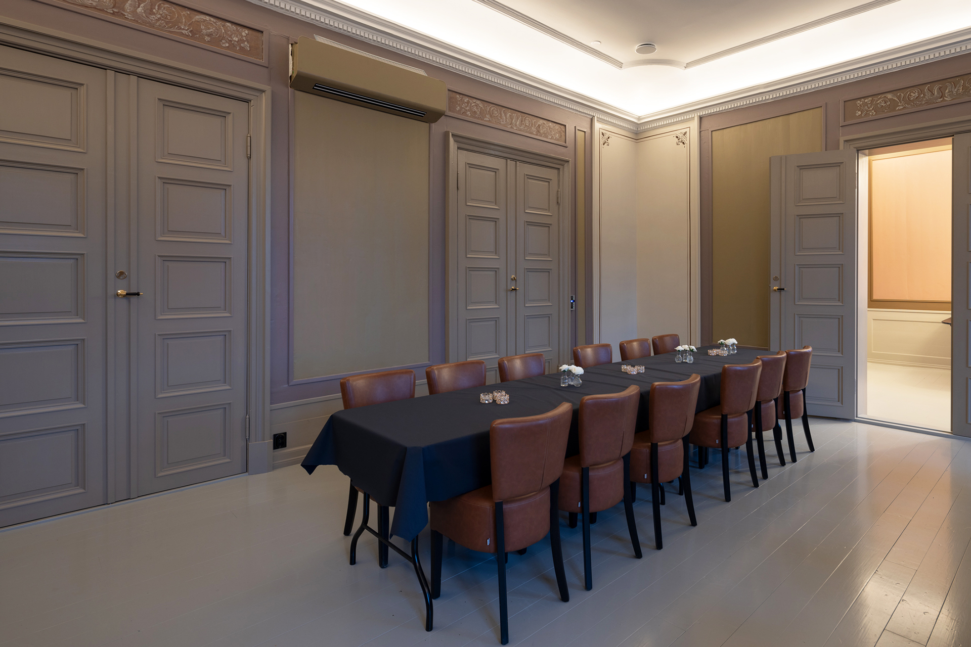 Et langbord med brune stoler står i et formelt møterom med panelvegger, lukkede dører og dempet takbelysning.