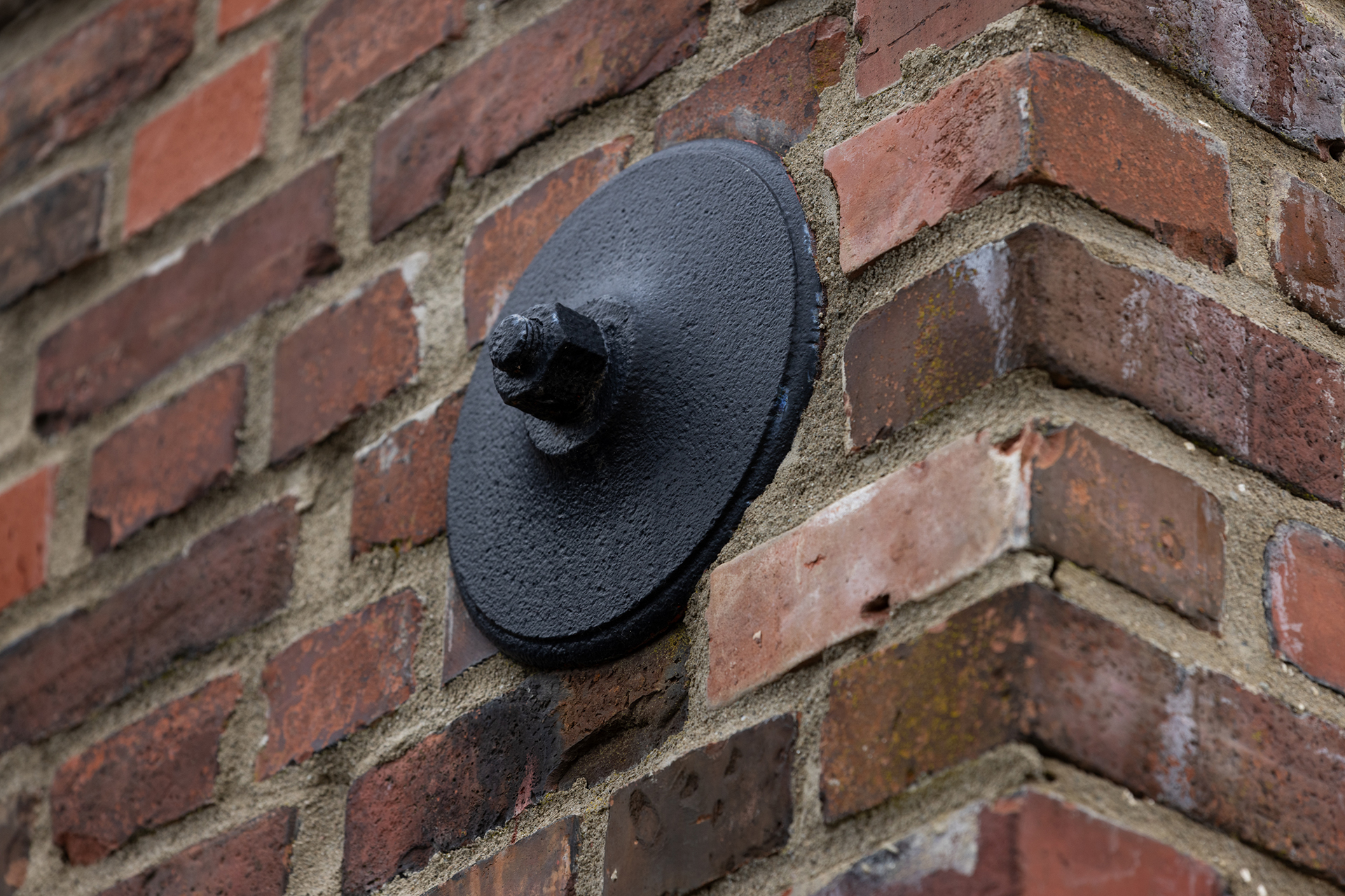 En stor, svart ankerplate av metall er boltet fast i hjørnet av en rød murvegg.