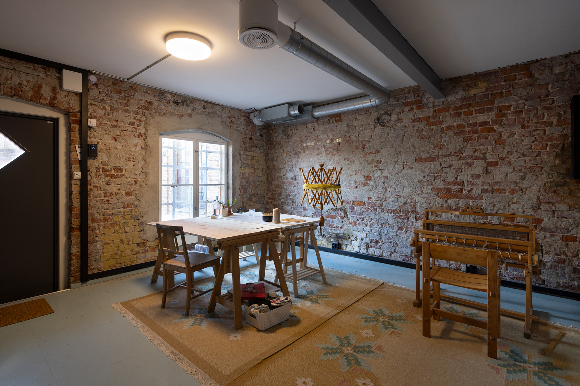 Sirkulærøkonomi i praksis 10 Et rom med synlige murvegger, trebord og -stoler, vevstoler og et mønstret teppe, opplyst av naturlig lys og taklys.